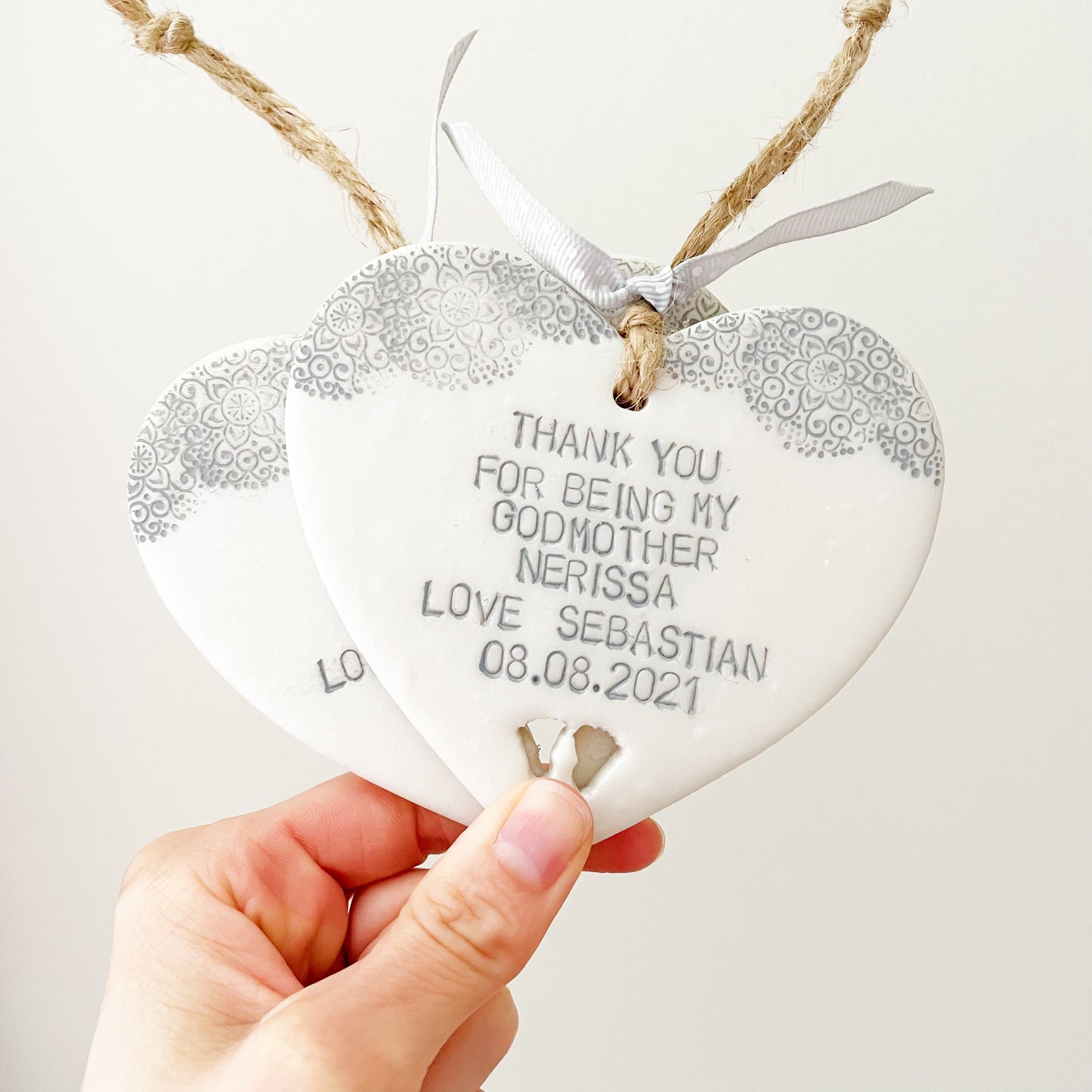 Personalised Godmother gift, 2 pearlised white clay hanging heart with baby feet cut out of the bottom and grey lace edge at the top of the heart, the heart is personalised with THANK YOU FOR BEING MY GODMOTHER NERISSA LOVE SEBASTIAN 08.08.2021