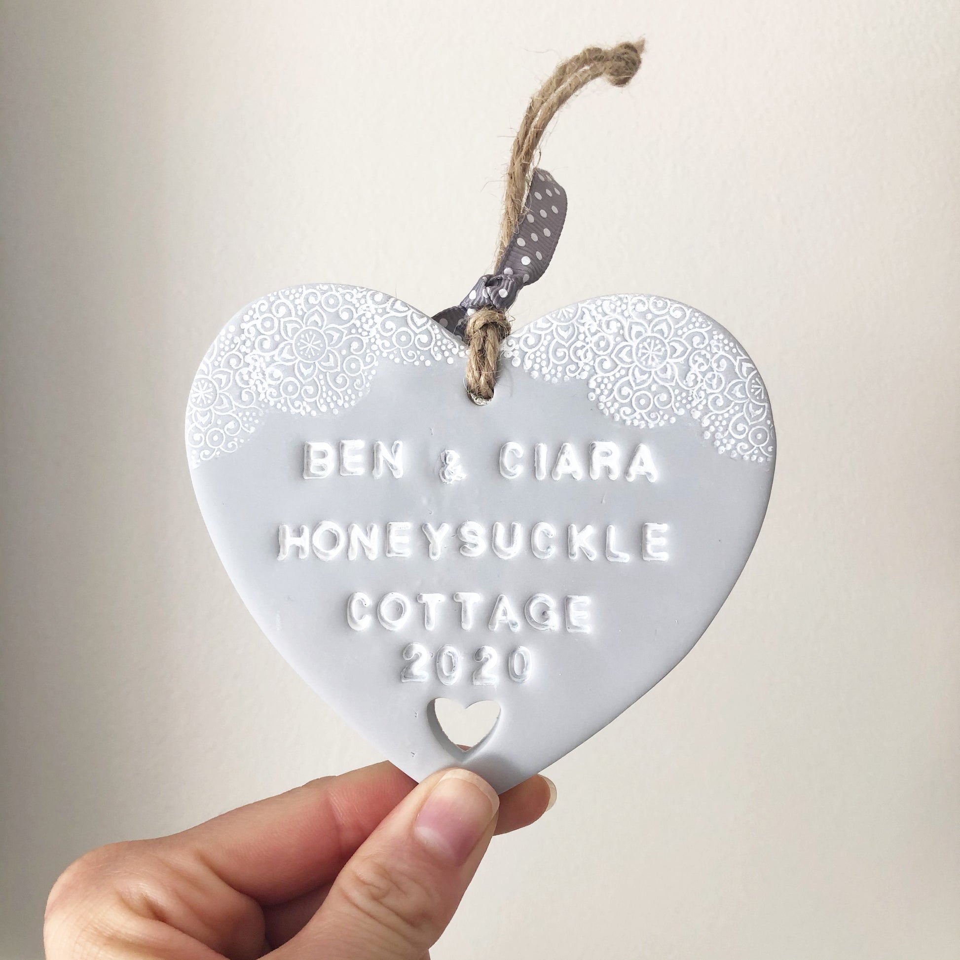 Personalised housewarming new home gift, grey clay heart with a white lace edge at the top of the heart and a heart cut out at the bottom with jute twine for hanging, the heart is personalised with BEN & CIARA HONEY SUCKLE COTTAGE 2020
