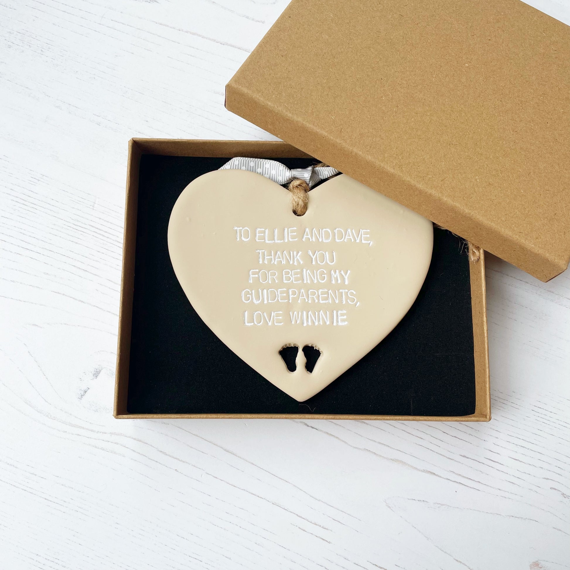 Personalised Guide parent gift, beige clay hanging heart with baby feet cut out of the bottom, the heart is personalised in white with TO ELLIE AND DAVE, THANK YOU FOR BEING MY GUIDEPARENTS, LOVE WINNIE