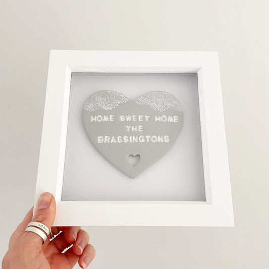 Personalised housewarming new home gift, grey clay heart with a white lace edge at the top of the heart and a heart cut out at the bottom in a white box frame, the heart is personalised with HOME SWEET HOME THE BRASSINGTONS