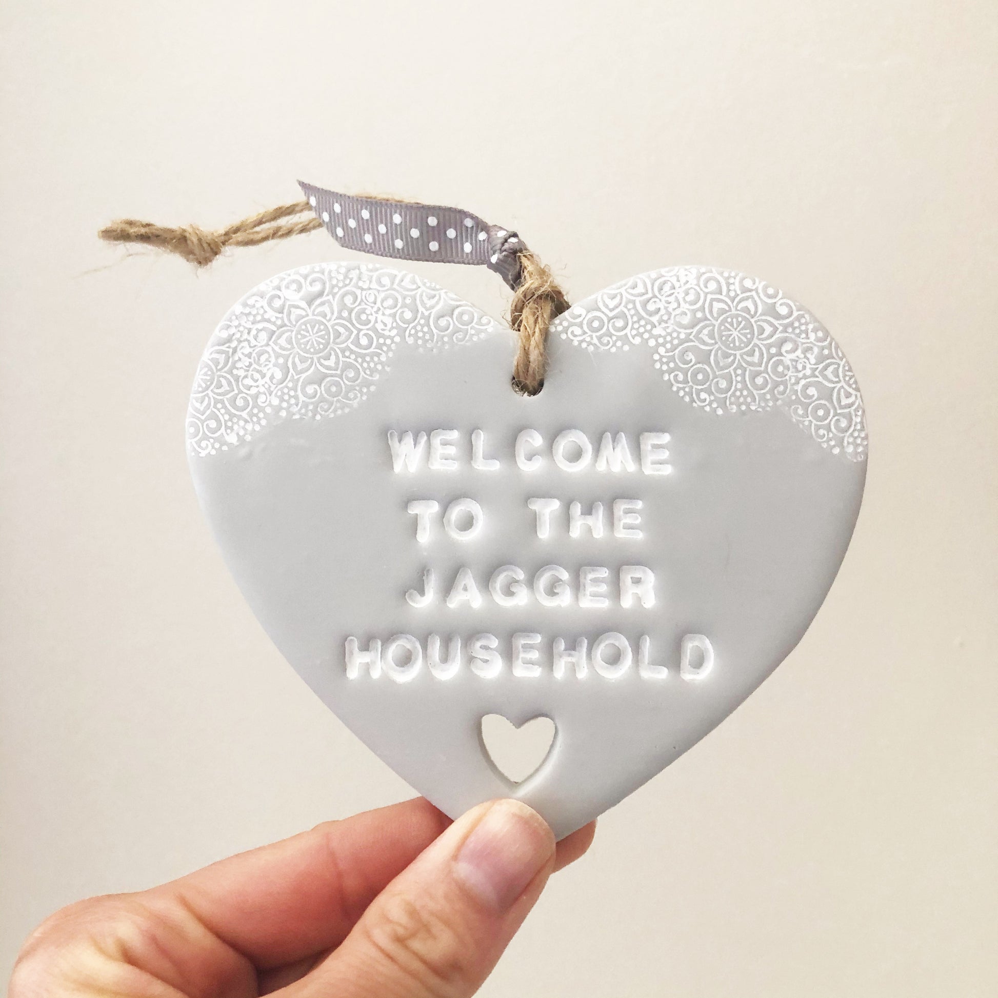 Personalised housewarming new home gift, grey clay heart with a white lace edge at the top of the heart and a heart cut out at the bottom with jute twine for hanging, the heart is personalised with WELCOME TO THE JAGGER HOUSEHOLD