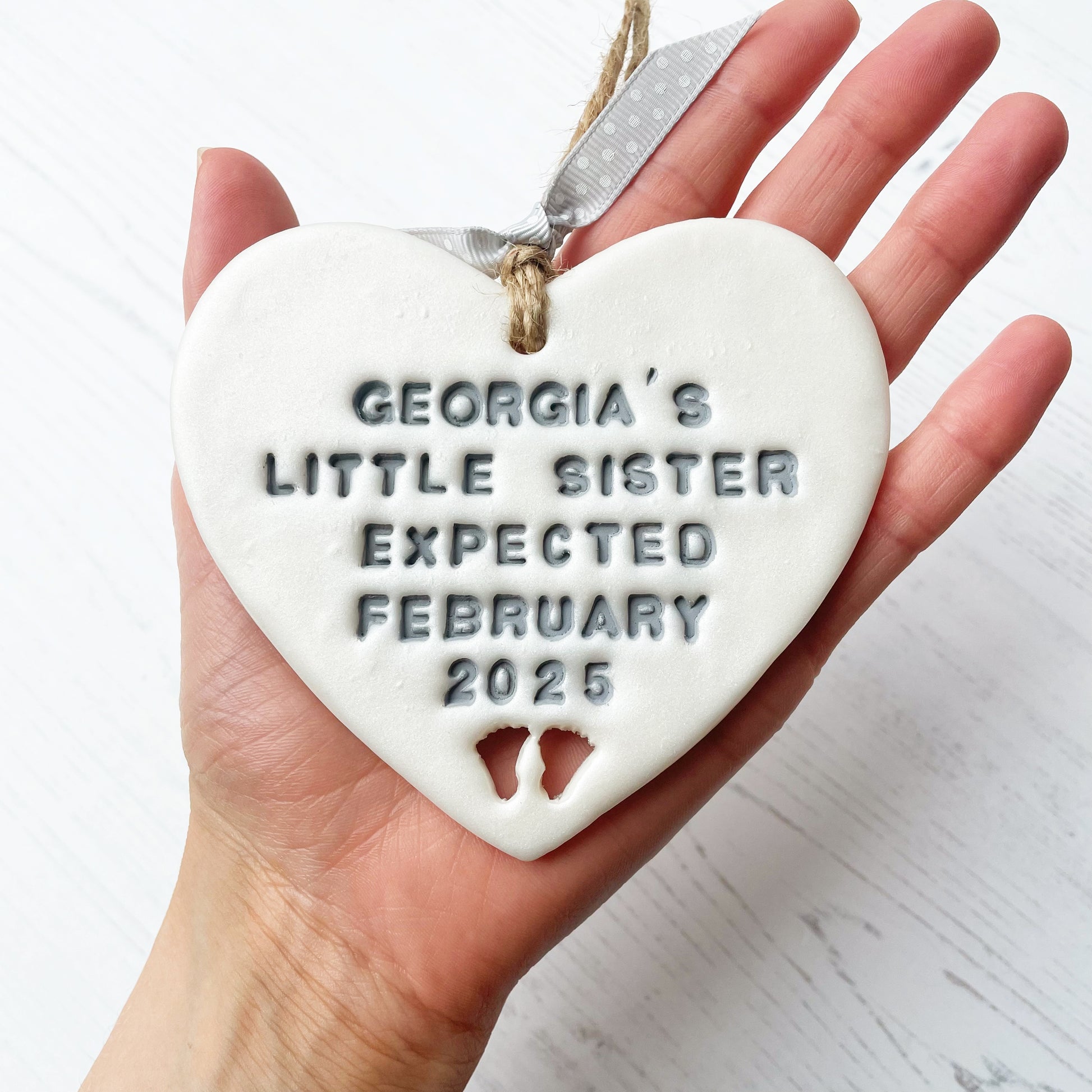 Personalised baby reveal gift, pearlised white clay heart with feet cut out of the bottom personalised in grey with GEORGIA’S LITTLE SISTER EXPECTED FEBRUARY 2025