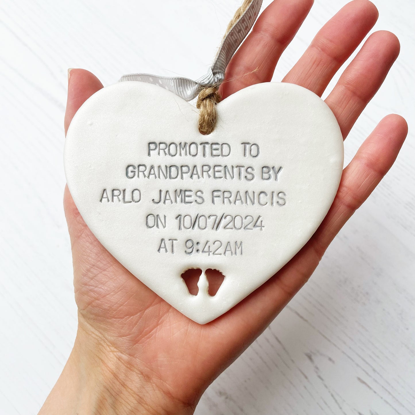 Pearlised white clay hanging heart with baby feet cut out of the bottom and grey personalisation, the heart is personalised with PROMOTED TO GRANDPARENTS BY ARLO JAMES FRANCIS ON 10/07/2024 AT 9:42 AM