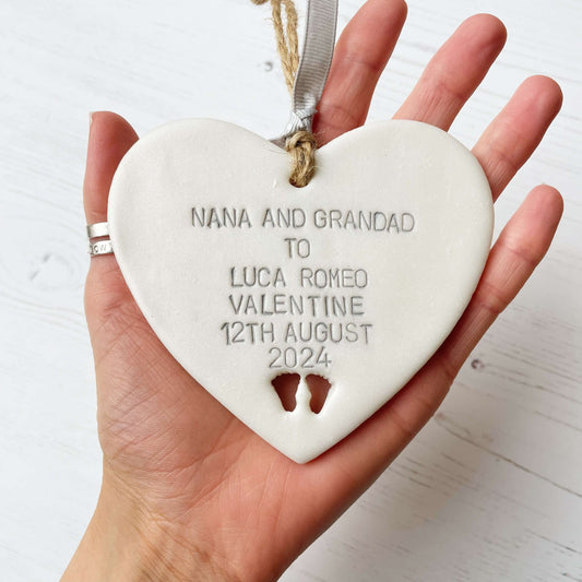 Pearlised white clay hanging heart with baby feet cut out of the bottom and grey personalisation, the heart is personalised with NANA AND GRANDAD TO LUCA ROMEO VALENTINE 12TH AUGUST 2024