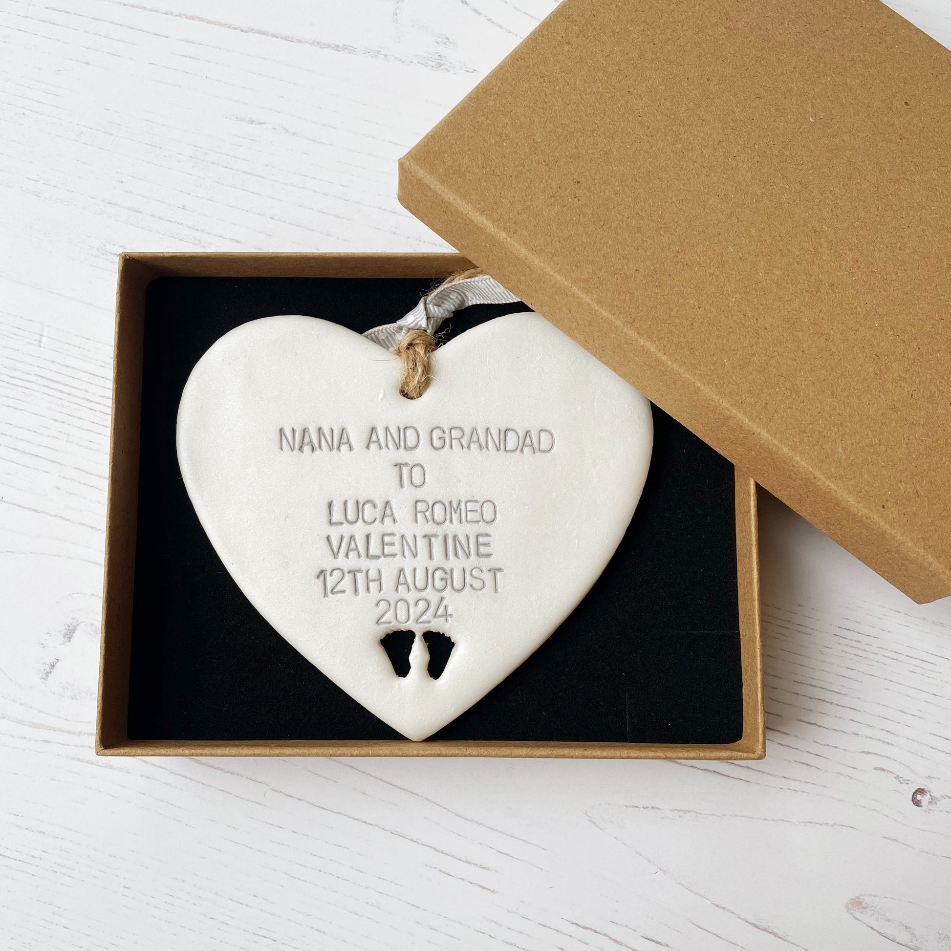 Pearlised white clay hanging heart with baby feet cut out of the bottom and grey personalisation, the heart is personalised with NANA AND GRANDAD TO LUCA ROMEO VALENTINE 12TH AUGUST 2024