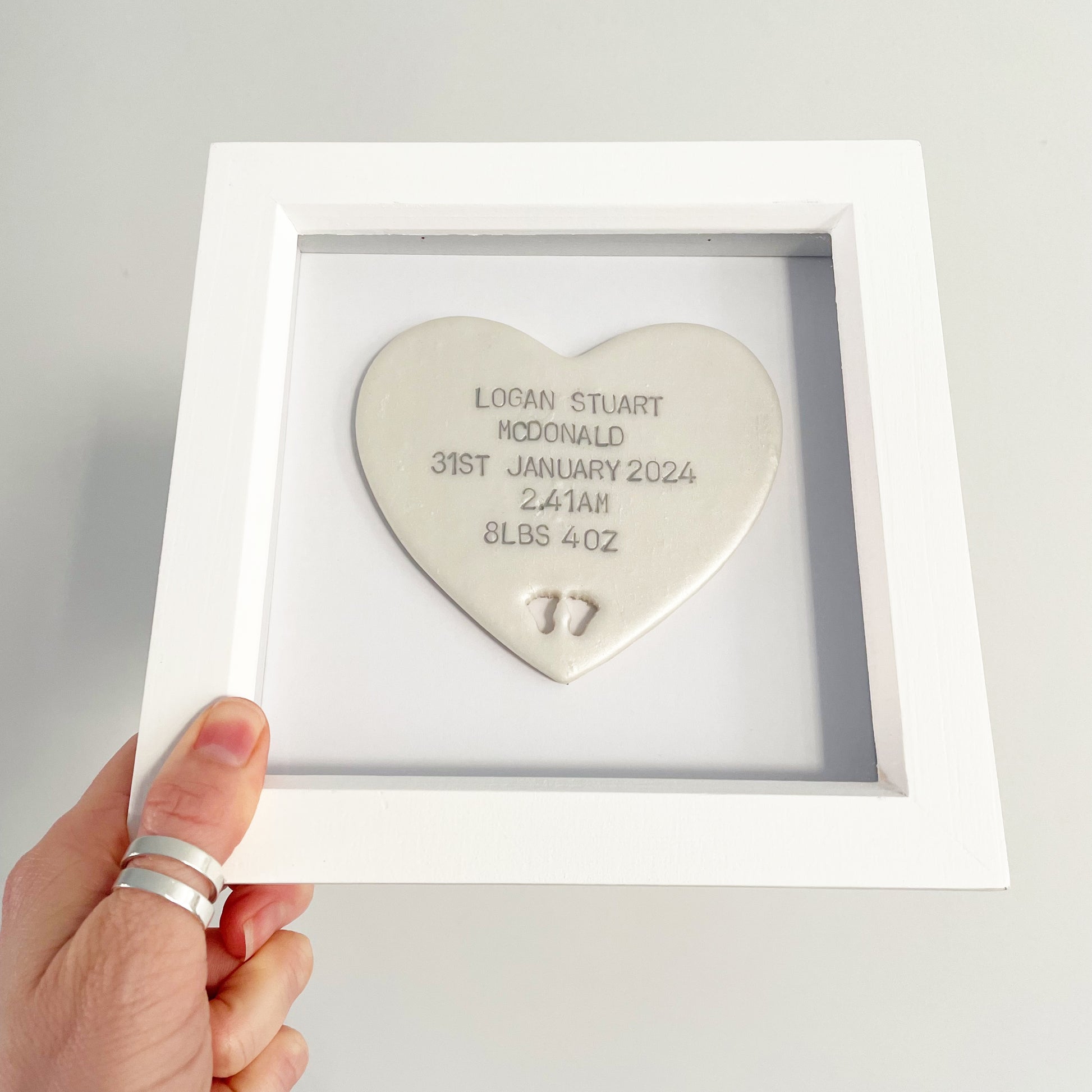 Personalised baby keepsake gift, pearlised white clay heart with baby feet cut out at the bottom of the heart and grey personalisation in a white box frame, the heart is personalised with the baby’s name, date of birth, weight and time