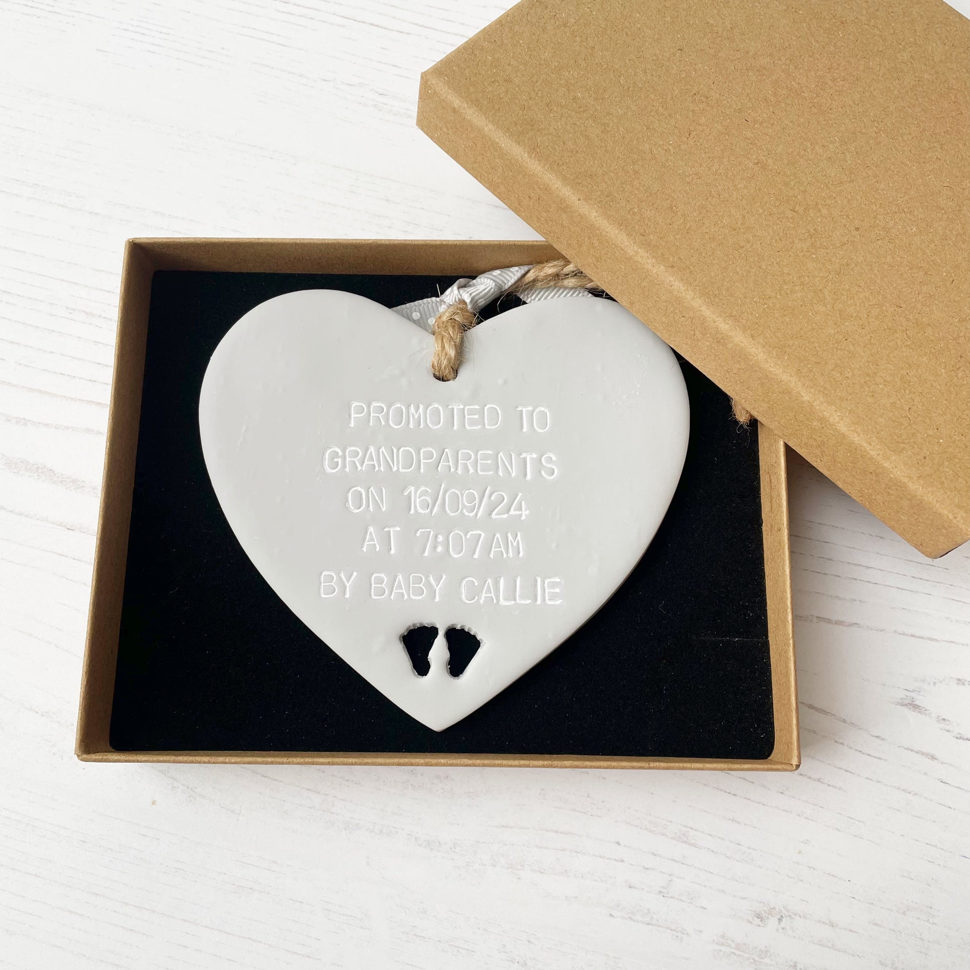 Grey clay hanging heart with baby feet cut out of the bottom and white personalisation, the heart is personalised with PROMOTED TO GRANDPARENTS ON 16/09/24 AT 7:07AM BY BABY CALLIE