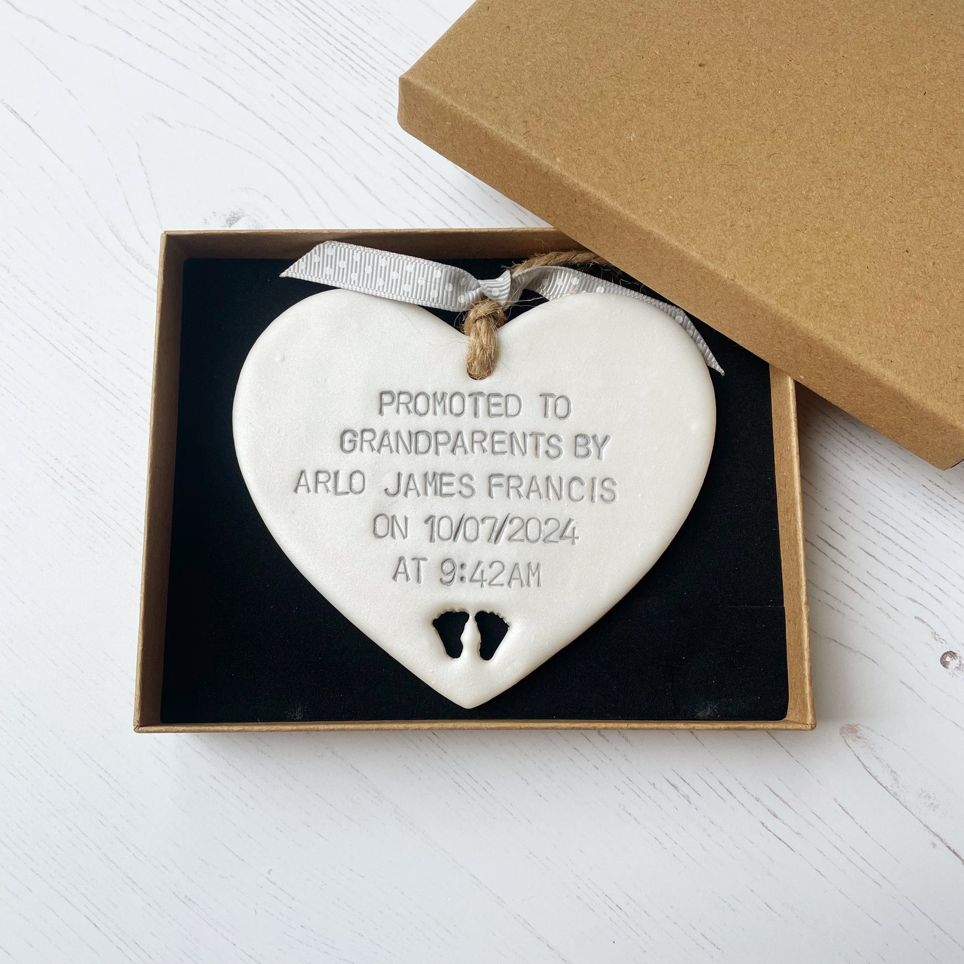 Pearlised white clay hanging heart with baby feet cut out of the bottom and grey personalisation, the heart is personalised with PROMOTED TO GRANDPARENTS BY ARLO JAMES FRANCIS ON 10/07/2024 AT 9:42 AM