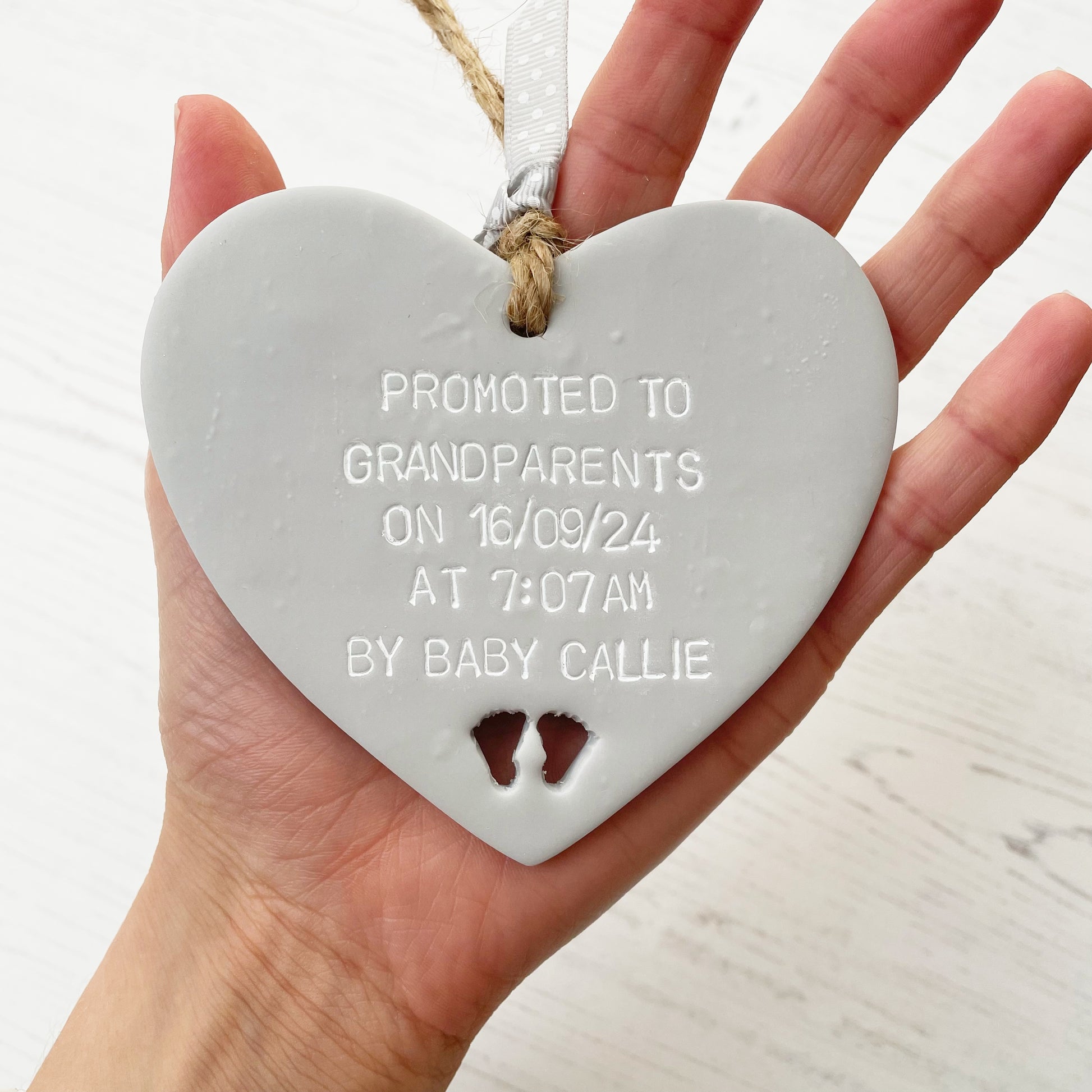 Grey clay hanging heart with baby feet cut out of the bottom and white personalisation, the heart is personalised with PROMOTED TO GRANDPARENTS ON 16/09/24 AT 7:07AM BY BABY CALLIE