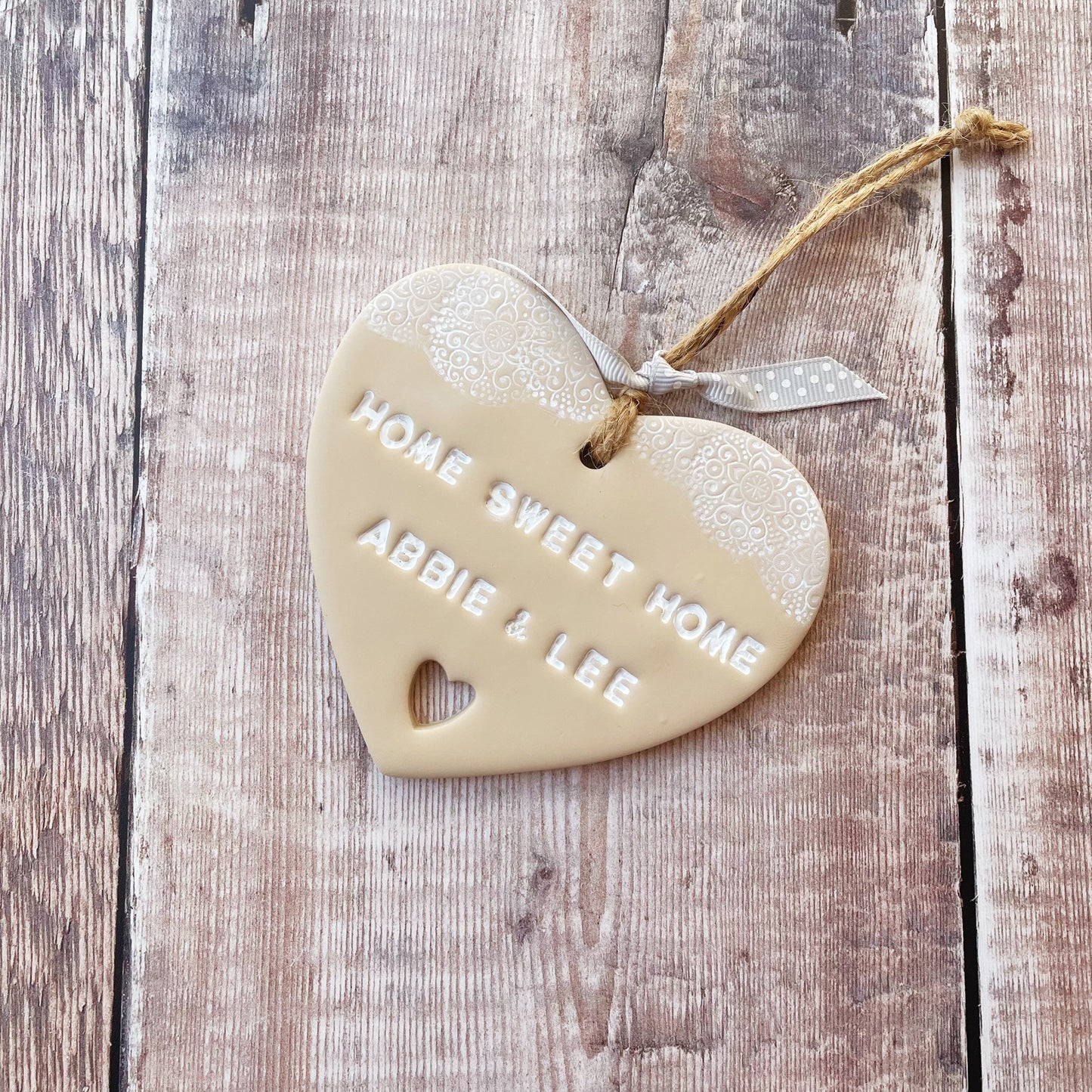 Personalised housewarming new home gift, beige clay heart with a white lace edge at the top of the heart and a heart cut out at the bottom with jute twine for hanging, the heart is personalised with HOME SWEET HOME ABBIE & LEE