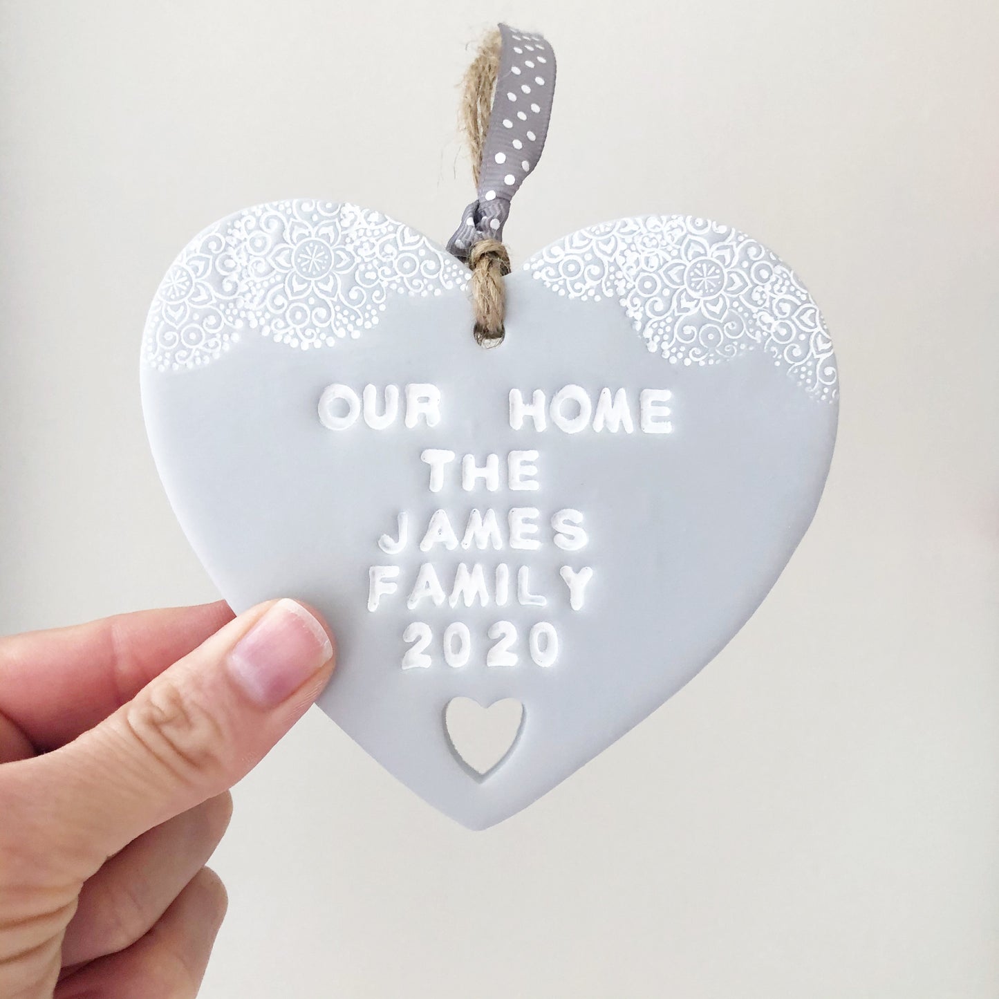 Personalised housewarming new home gift, grey clay heart with a white lace edge at the top of the heart and a heart cut out at the bottom with jute twine for hanging, the heart is personalised with OUR HOME THE JAMES FAMILY 2020