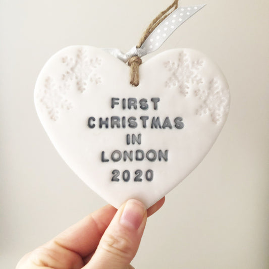 Personalised first Christmas in our new home heart ornament, pearlised white clay with FIRST CHRISTMAS IN LONDON 2020 painted grey, decorated with 2 iridescent glitter snowflakes on either side of the top of the heart