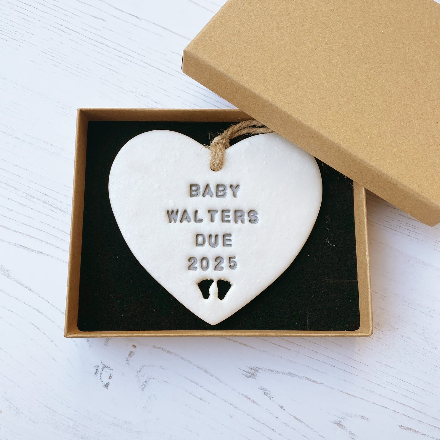 Personalised pregnancy reveal sign keepsake, pearlised white clay hanging heart with baby feet cut out at the bottom, the heart is personalised in grey with BABY WALTERS DUE 2025