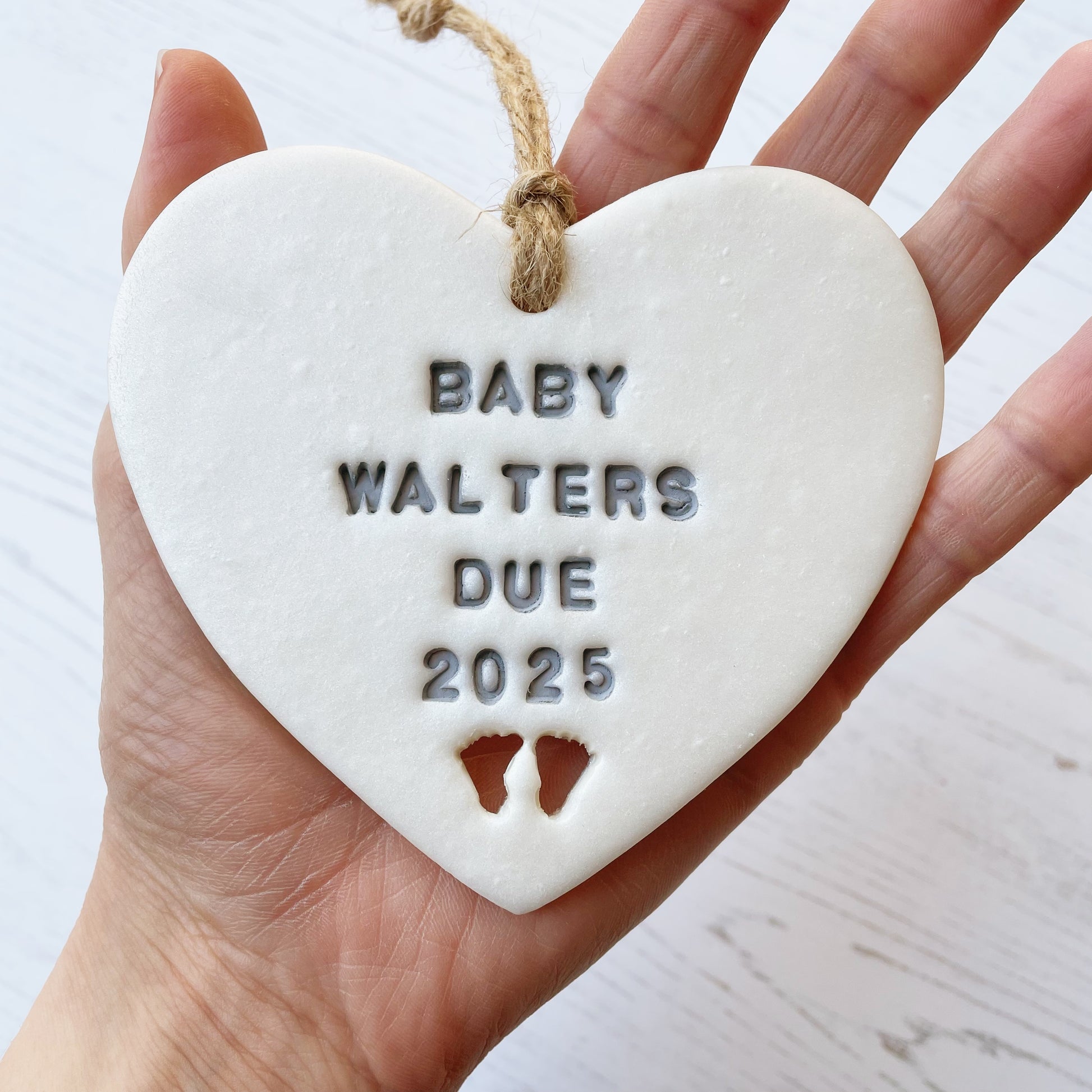 Personalised pregnancy reveal sign keepsake, pearlised white clay hanging heart with baby feet cut out at the bottom, the heart is personalised in grey with BABY WALTERS DUE 2025