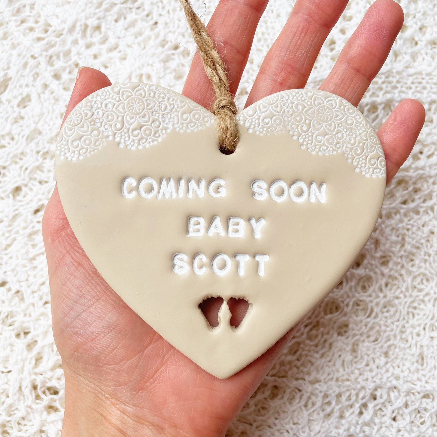 Personalised pregnancy reveal sign keepsake, beige clay hanging heart with a white lace edge at the top of the heart and baby feet cut out at the bottom, the heart is personalised with COMING SOON BABY SCOTT