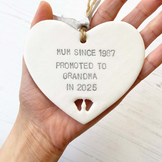 Pearlised white clay hanging heart with baby feet cut out of the bottom and grey personalisation, the heart is personalised with MUM SINCE 1987 PROMOTED TO GRANDMA IN 2025