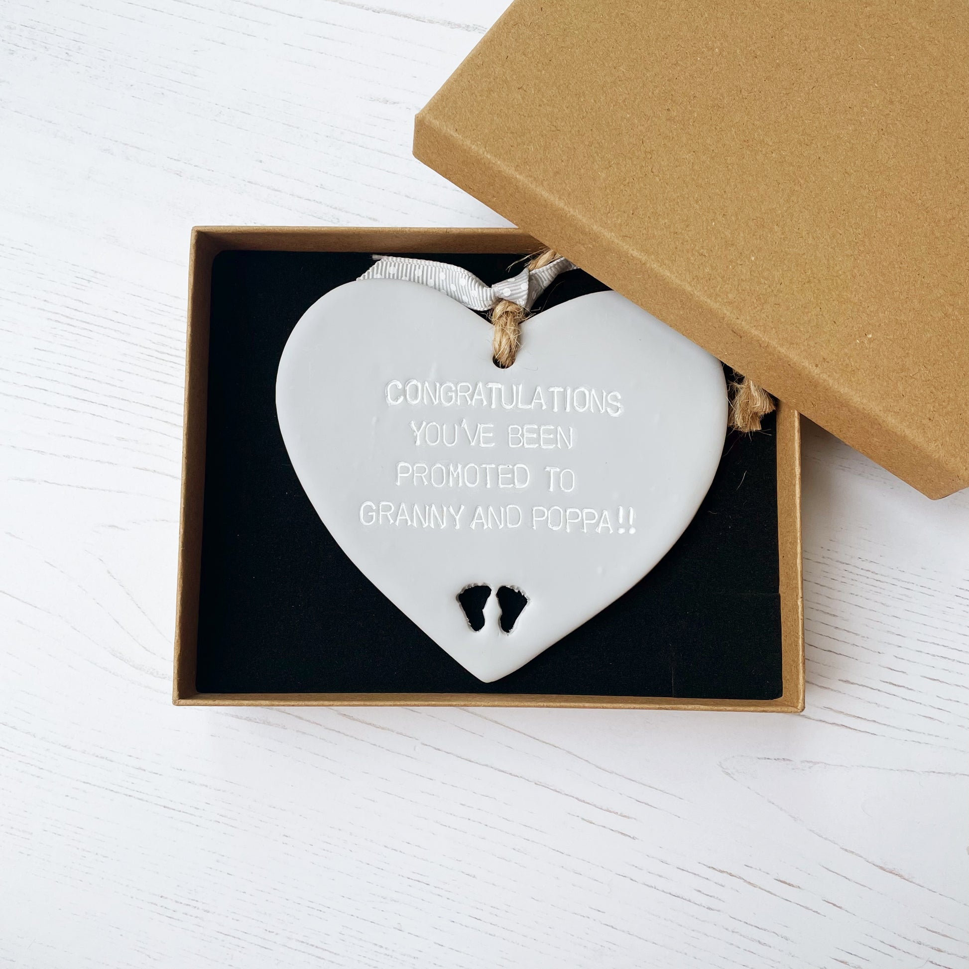 Grey clay hanging heart with baby feet cut out of the bottom and white personalisation, the heart is personalised with CONGRATULATIONS YOU’CE BEEN PROMOTED TO GRANNY AND POPPA!!