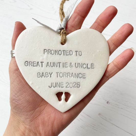 Pearlised white clay hanging heart with baby feet cut out of the bottom and grey personalisation, the heart is personalised with PROMOTED TO GREAT AUNTIE & UNCLE BABY TORRANCE JUNE 2025