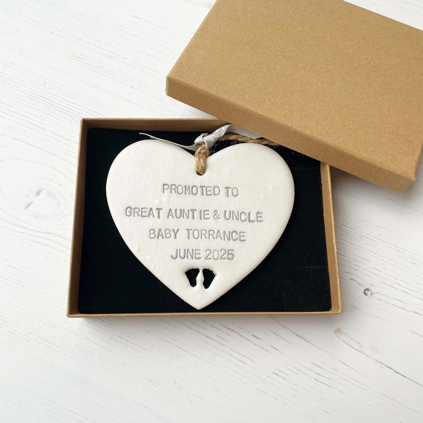 Pearlised white clay hanging heart with baby feet cut out of the bottom and grey personalisation, the heart is personalised with PROMOTED TO GREAT AUNTIE & UNCLE BABY TORRANCE JUNE 2025