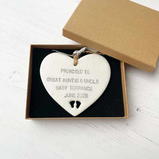 Pearlised white clay hanging heart with baby feet cut out of the bottom and grey personalisation, the heart is personalised with PROMOTED TO GREAT AUNTIE & UNCLE BABY TORRANCE JUNE 2025