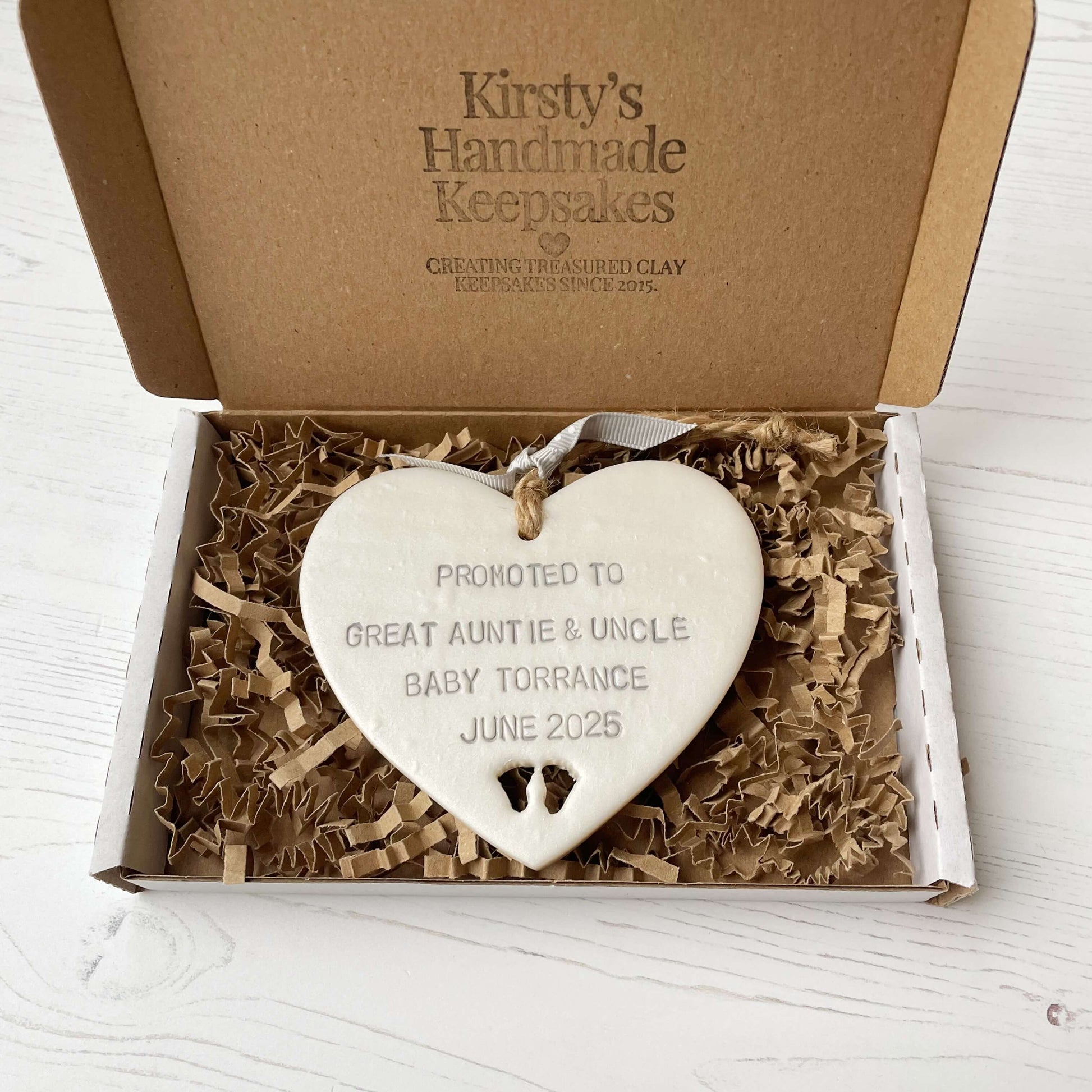 Pearlised white clay hanging heart with baby feet cut out of the bottom and grey personalisation, the heart is personalised with PROMOTED TO GREAT AUNTIE & UNCLE BABY TORRANCE JUNE 2025