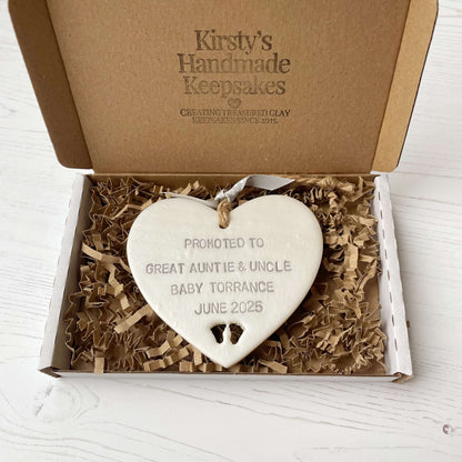 Pearlised white clay hanging heart with baby feet cut out of the bottom and grey personalisation, the heart is personalised with PROMOTED TO GREAT AUNTIE & UNCLE BABY TORRANCE JUNE 2025