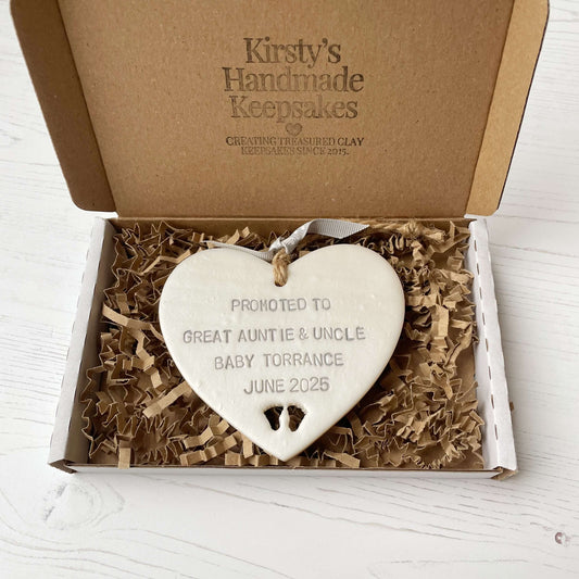 Pearlised white clay hanging heart with baby feet cut out of the bottom and grey personalisation, the heart is personalised with PROMOTED TO GREAT AUNTIE & UNCLE BABY TORRANCE JUNE 2025