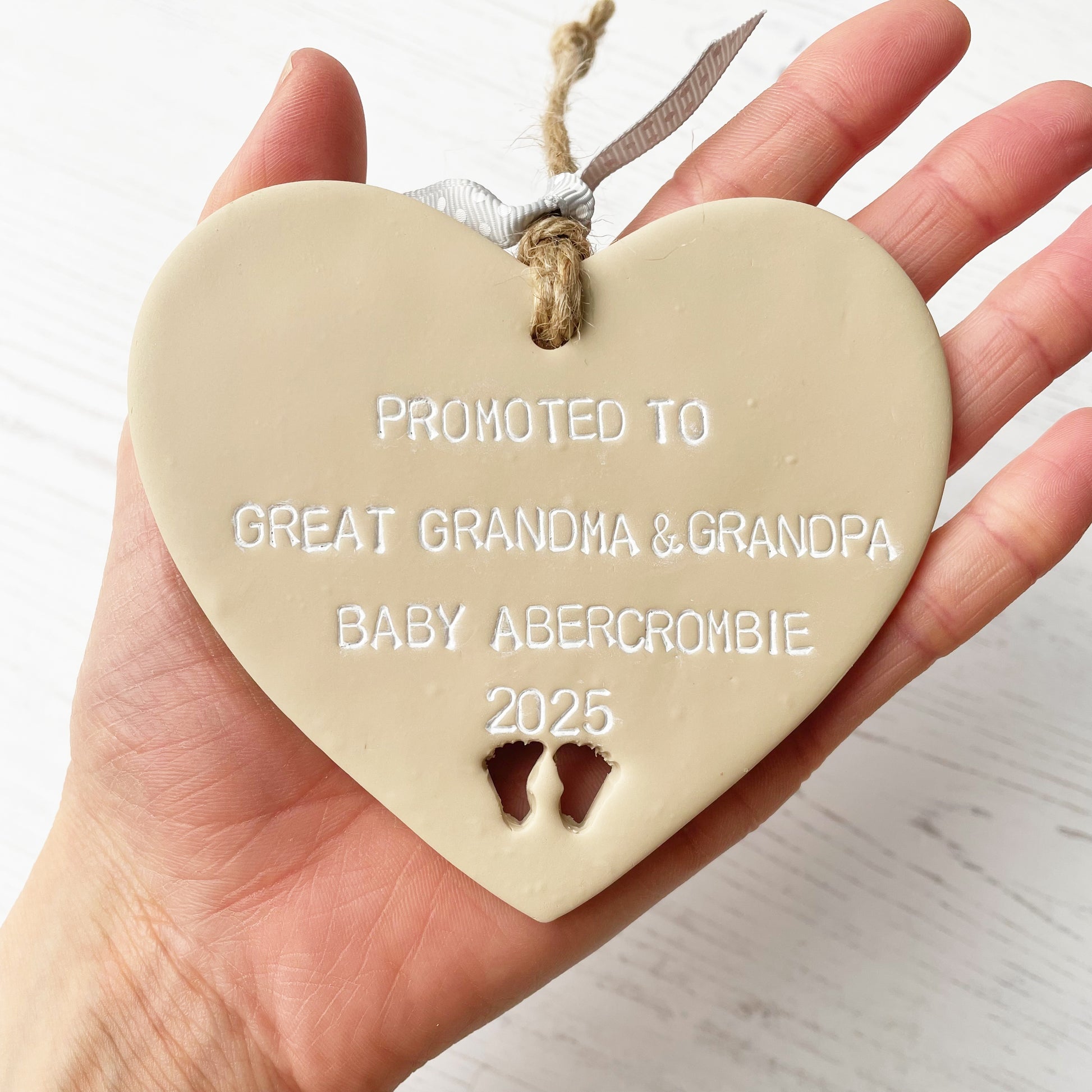 Beige clay hanging heart with baby feet cut out of the bottom and white personalisation, the heart is personalised with PROMOTED TO GREAT GRANDMA & GRANDPA BABY ABERCROMBIE 2025