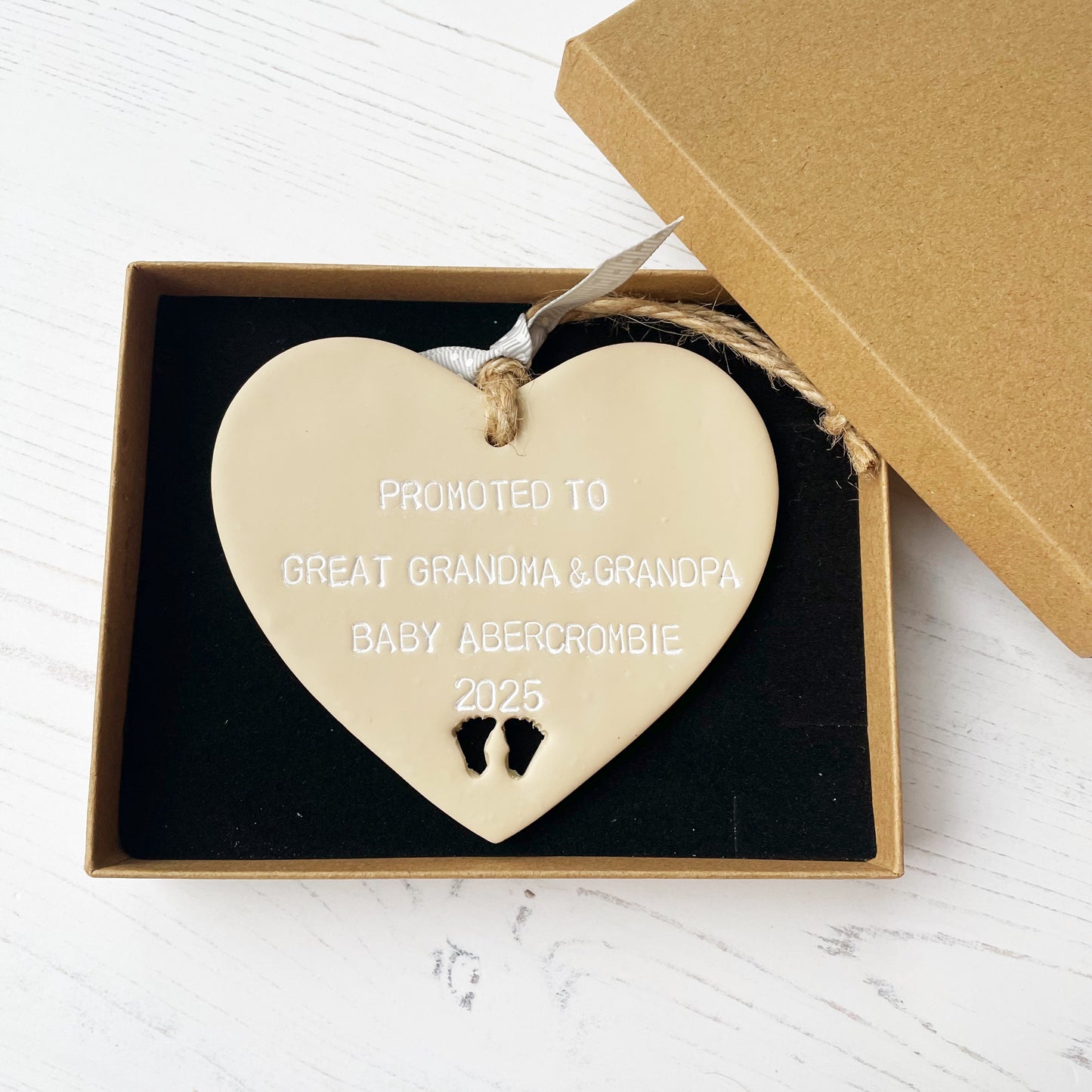 Beige clay hanging heart with baby feet cut out of the bottom and white personalisation, the heart is personalised with PROMOTED TO GREAT GRANDMA & GRANDPA BABY ABERCROMBIE 2025