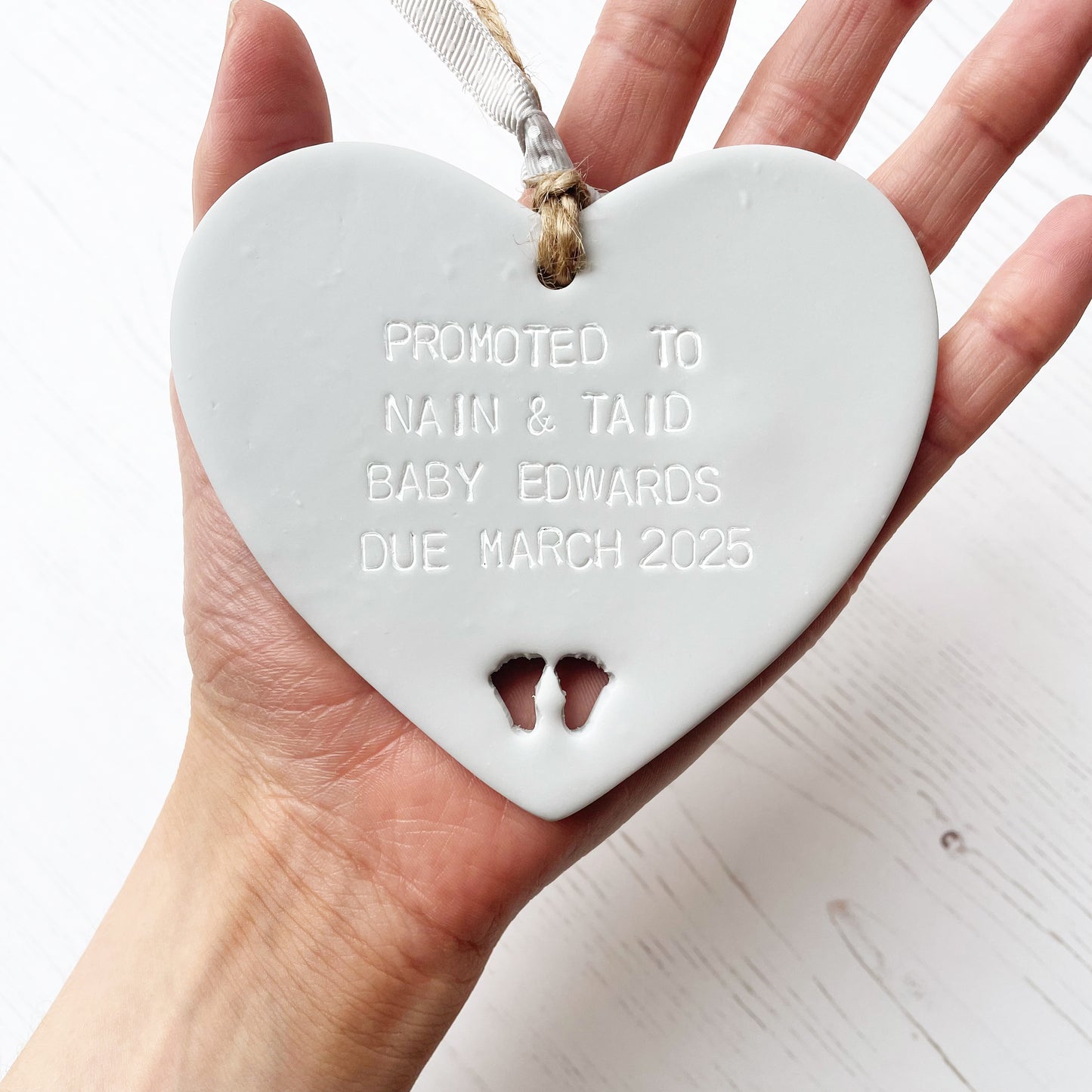 Grey clay hanging promoted to Grandparents heart with baby feet cut out of the bottom and white personalisation, the heart is personalised with PROMOTED TO NAIN & TAID BABY EDWARDS DUE MARCH 2025