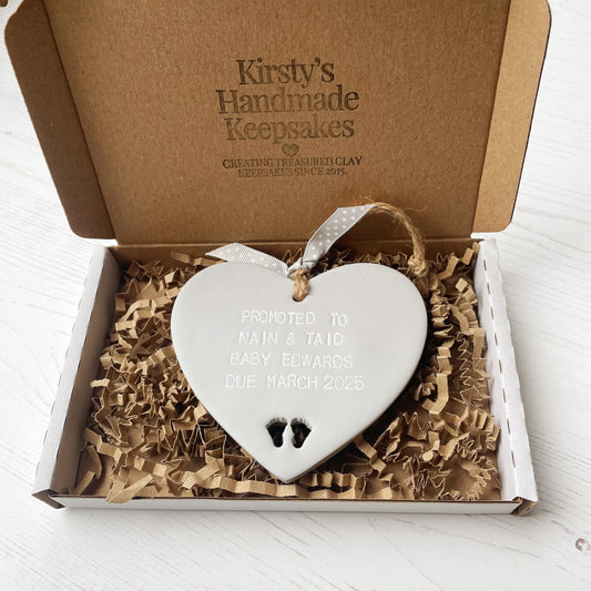 Grey clay hanging promoted to Grandparents heart with baby feet cut out of the bottom and white personalisation, the heart is personalised with PROMOTED TO NAIN & TAID BABY EDWARDS DUE MARCH 2025