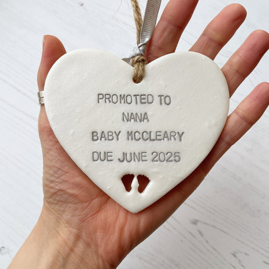 Pearlised white clay hanging heart with baby feet cut out of the bottom and grey personalisation, the heart is personalised with PROMOTED TO NANA BABY MCCLEARY DUE JUNE 2025