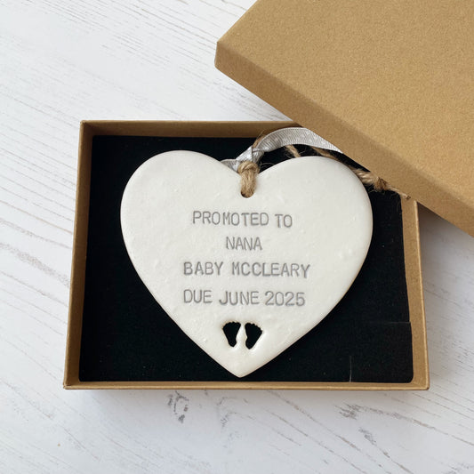 Pearlised white clay hanging heart with baby feet cut out of the bottom and grey personalisation, the heart is personalised with PROMOTED TO NANA BABY MCCLEARY DUE JUNE 2025