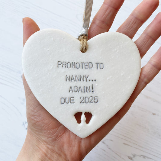 Pearlised white clay hanging heart with baby feet cut out of the bottom and grey personalisation, the heart is personalised with PROMOTED TO NANNY… AGAIN! DUE 2025