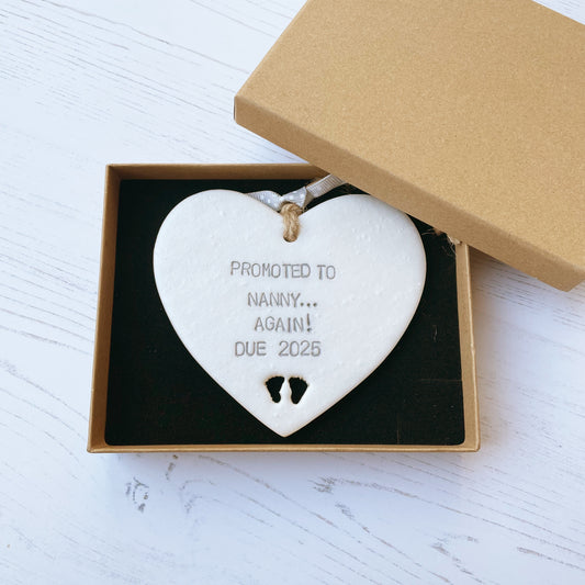 Pearlised white clay hanging heart with baby feet cut out of the bottom and grey personalisation, the heart is personalised with PROMOTED TO NANNY… AGAIN! DUE 2025