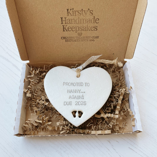 Pearlised white clay hanging heart with baby feet cut out of the bottom and grey personalisation, the heart is personalised with PROMOTED TO NANNY… AGAIN! DUE 2025