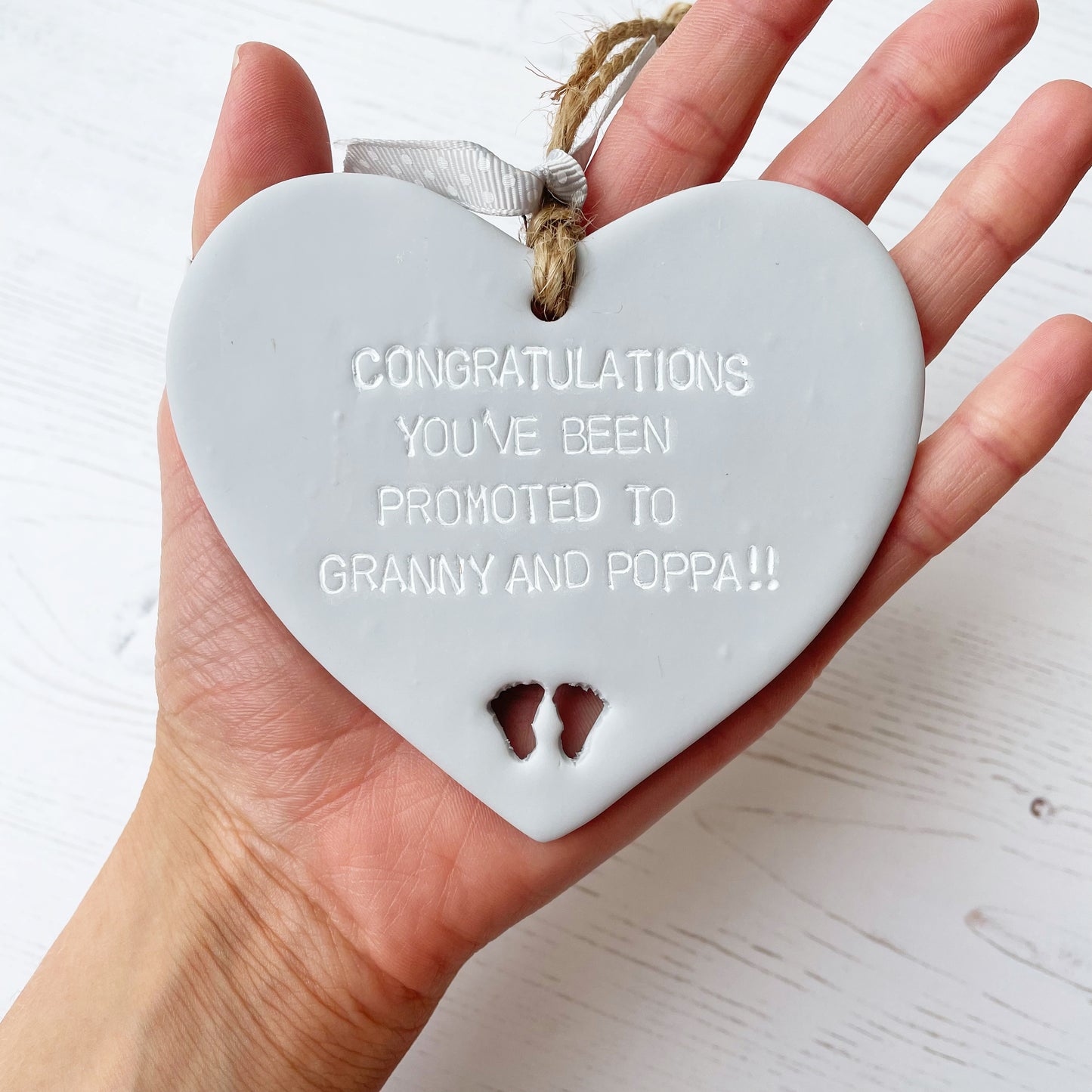 Grey clay hanging heart with baby feet cut out of the bottom and white personalisation, the heart is personalised with CONGRATULATIONS YOU’CE BEEN PROMOTED TO GRANNY AND POPPA!!