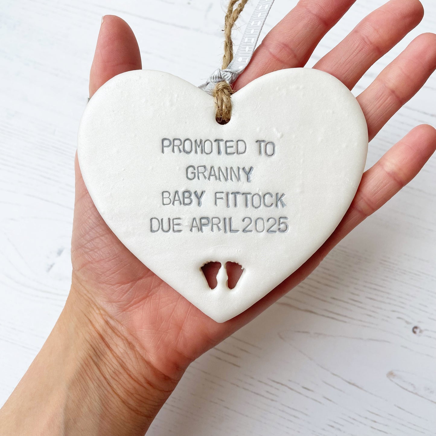 Pearlised white clay hanging heart with baby feet cut out of the bottom and grey personalisation, the heart is personalised with PROMOTED TO GRANNY BABY FITTOCK DUE APRIL 2025