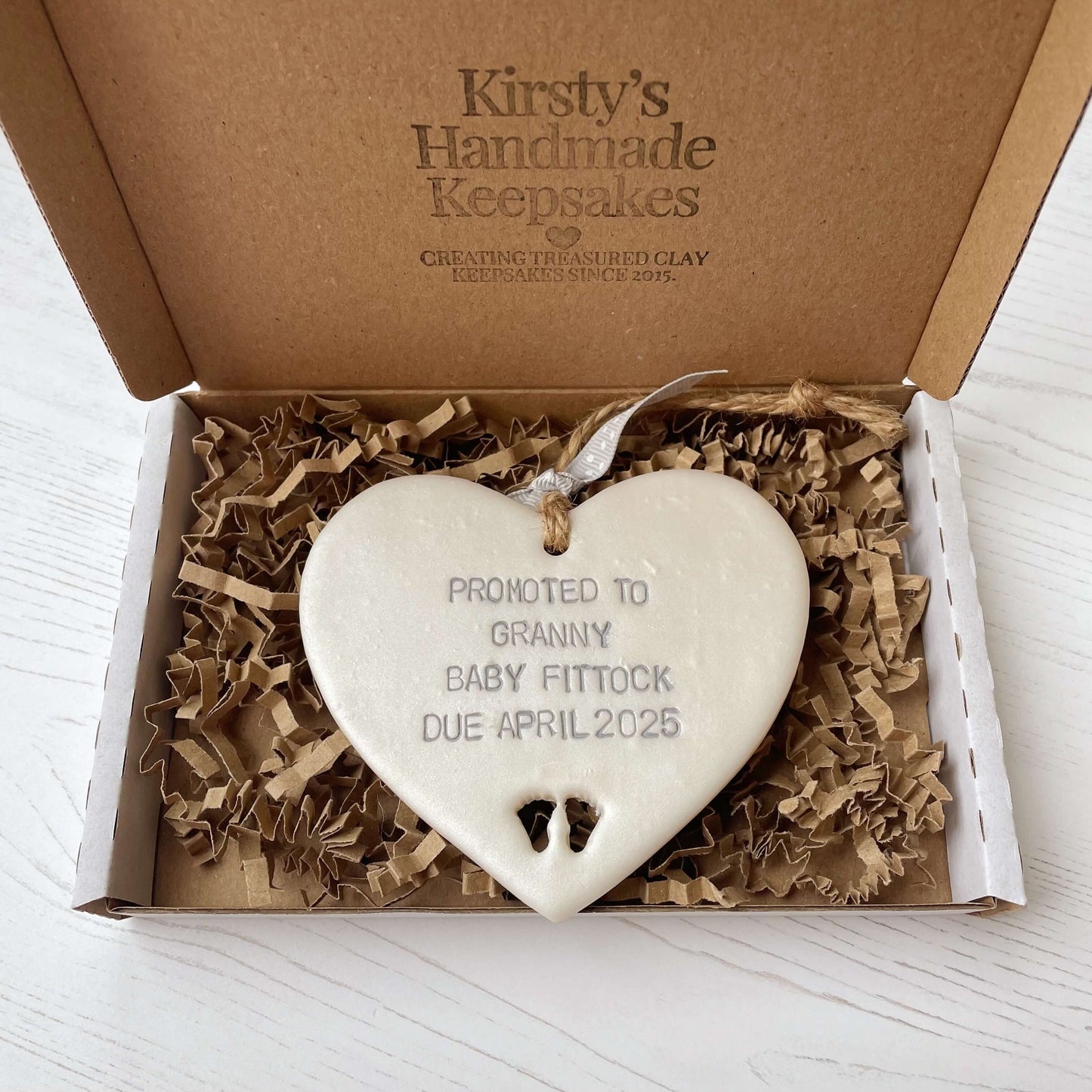 Pearlised white clay hanging heart with baby feet cut out of the bottom and grey personalisation, the heart is personalised with PROMOTED TO GRANNY BABY FITTOCK DUE APRIL 2025