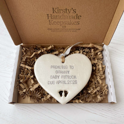 Pearlised white clay hanging heart with baby feet cut out of the bottom and grey personalisation, the heart is personalised with PROMOTED TO GRANNY BABY FITTOCK DUE APRIL 2025