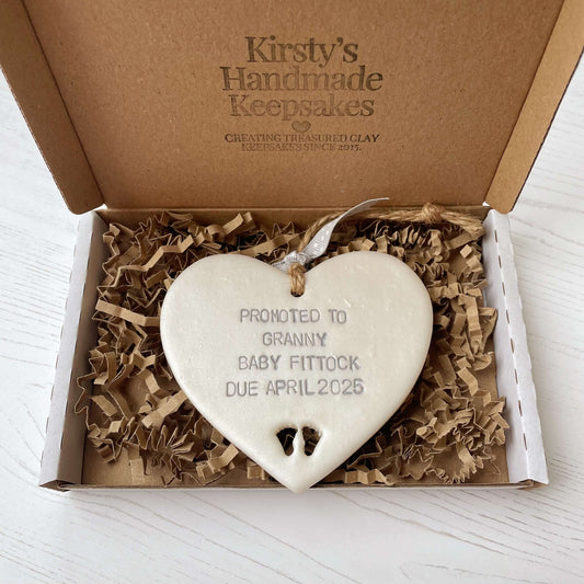 Pearlised white clay hanging heart with baby feet cut out of the bottom and grey personalisation, the heart is personalised with PROMOTED TO GRANNY BABY FITTOCK DUE APRIL 2025