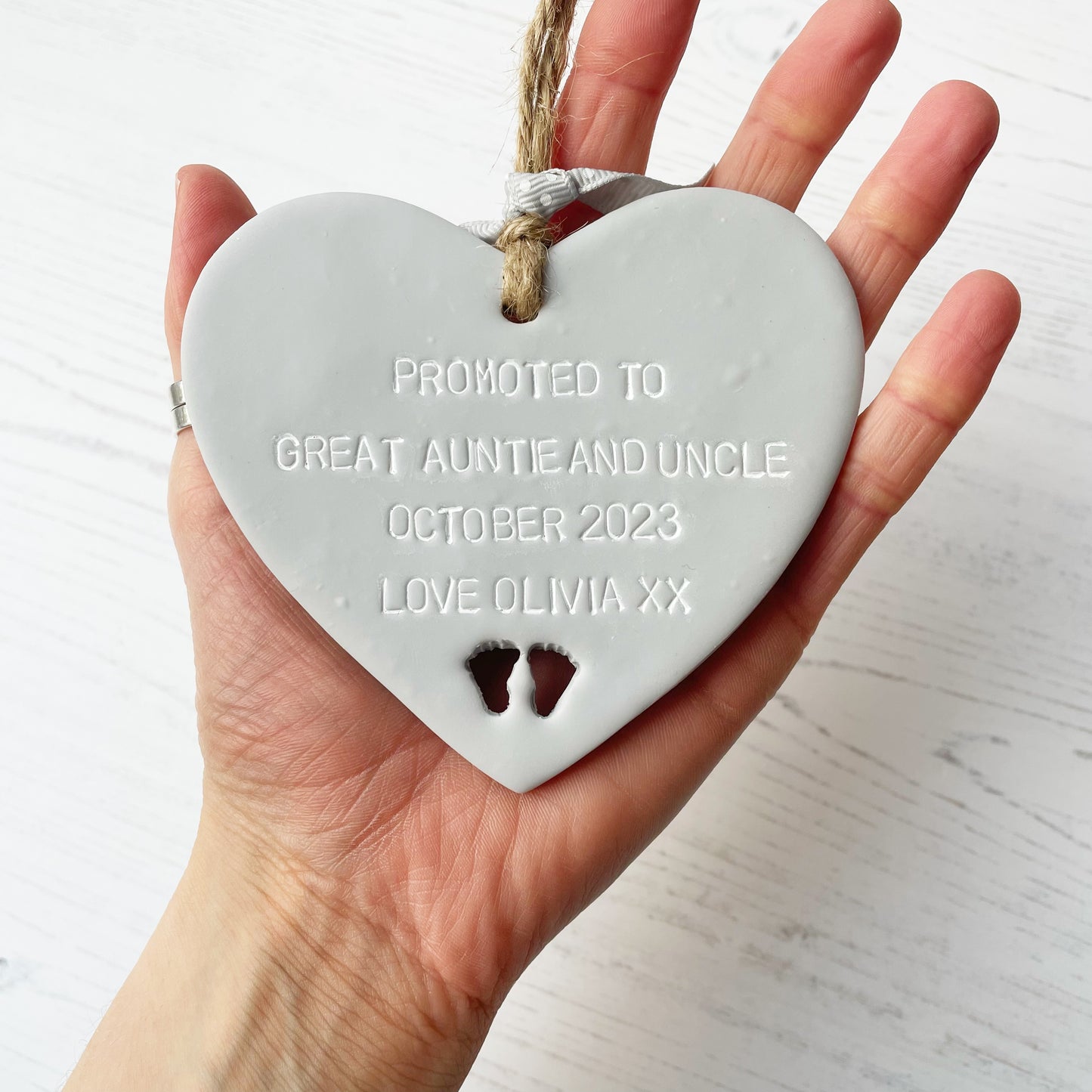 Grey clay hanging heart with baby feet cut out of the bottom and white personalisation, the heart is personalised with PROMOTED TO GREAT AUNTIE AND UNCLE OCTOBER 2023 LOVE OLIVIA XX