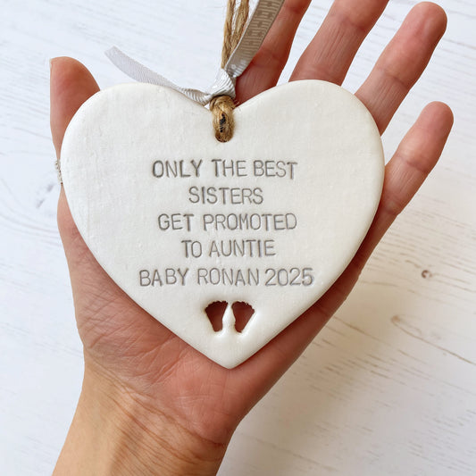 Pearlised white clay hanging heart with baby feet cut out of the bottom and grey personalisation, the heart is personalised with ONLY THE BEST SISTERS GET PROMOTED TO AUNTIE BABY RONAN 2025