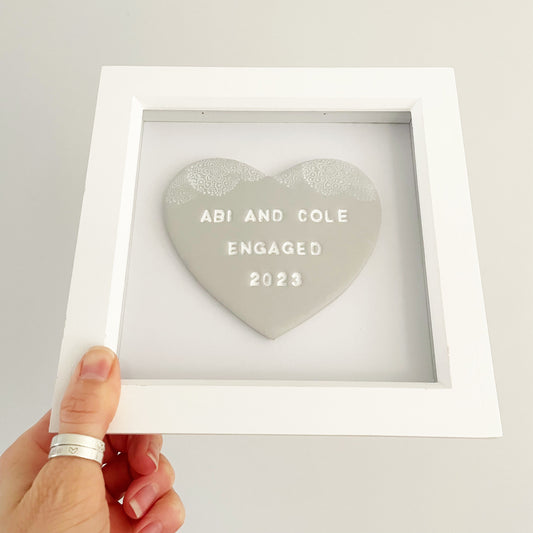 Personalised framed engagement gift, grey clay heart with a white lace edge at the top of the heart in a white box frame, the heart is personalised with ABI AND COLE ENGAGED 2023