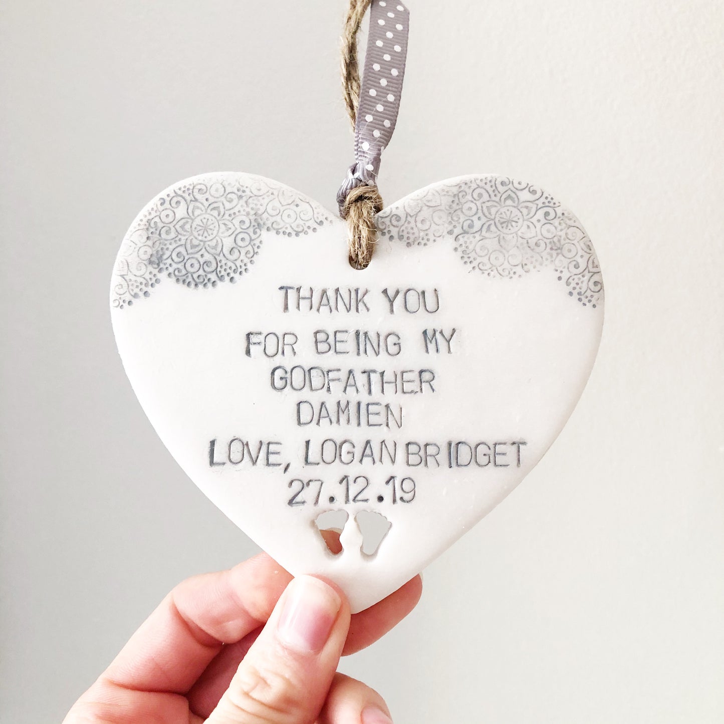 Personalised Godfather gift, pearlised white clay hanging heart with baby feet cut out of the bottom and a grey lace edge top, the heart is personalised with THANK YOU FOR BEING MY GODFATHER DAMIEN LOVE, LOGAN BRIDGET 27.12.19