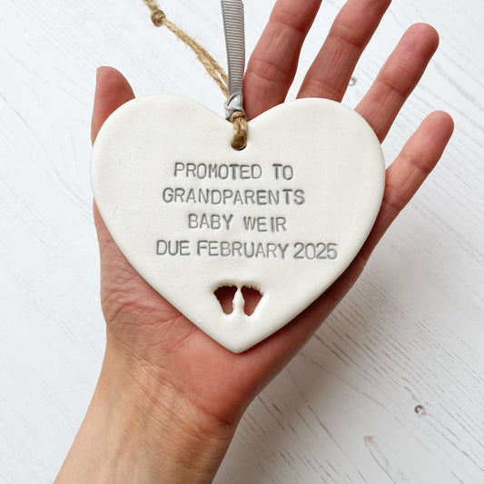Pearlised white clay hanging heart with baby feet cut out of the bottom and grey personalisation, the heart is personalised with PROMOTED TO GRANDPARENTS. BABY WEIR DUE FEBRUARY 2025