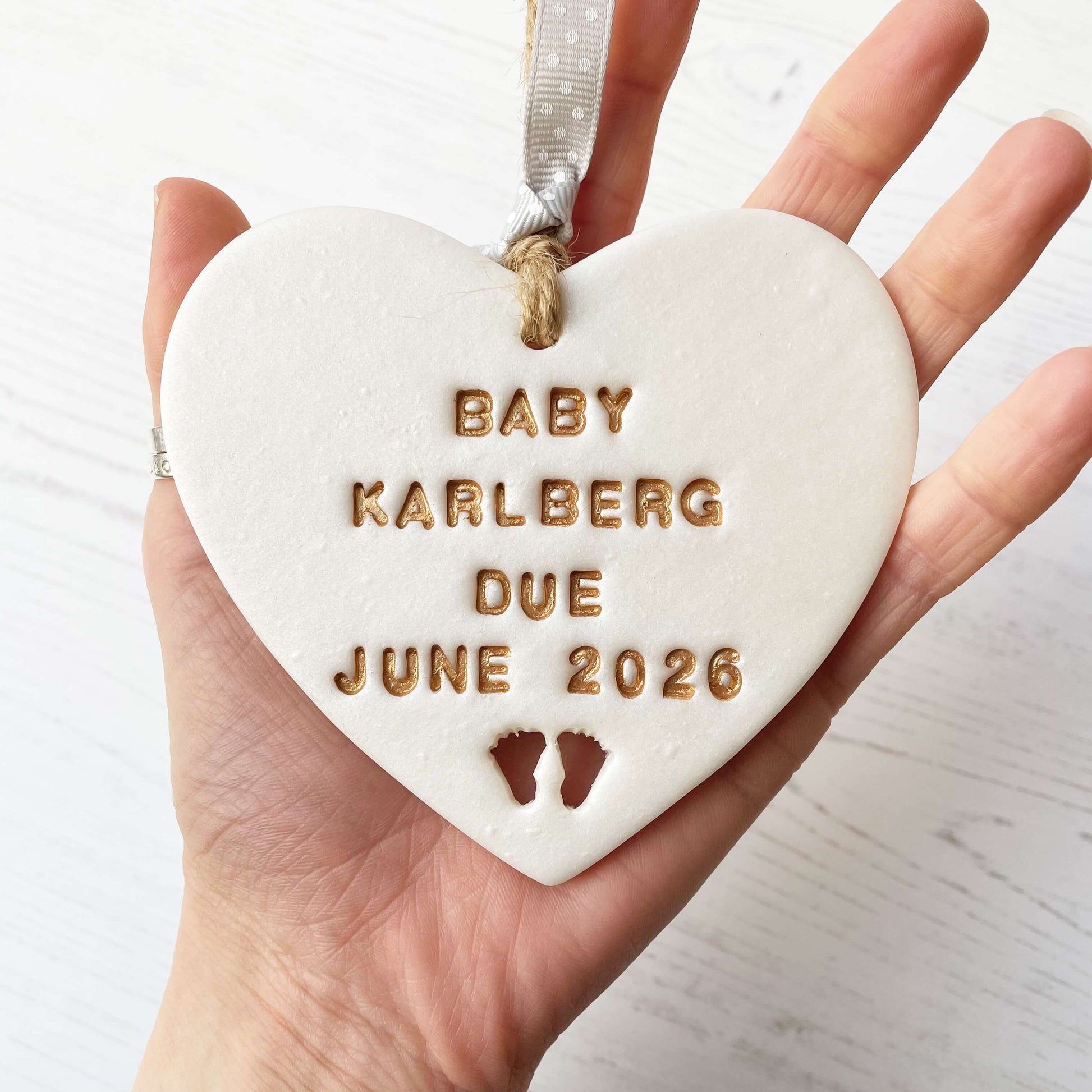 Personalised pregnancy reveal sign keepsake, pearlised white clay hanging heart with baby feet cut out at the bottom, the heart is personalised in gold with BABY KARLBERG DUE JUNE 2026