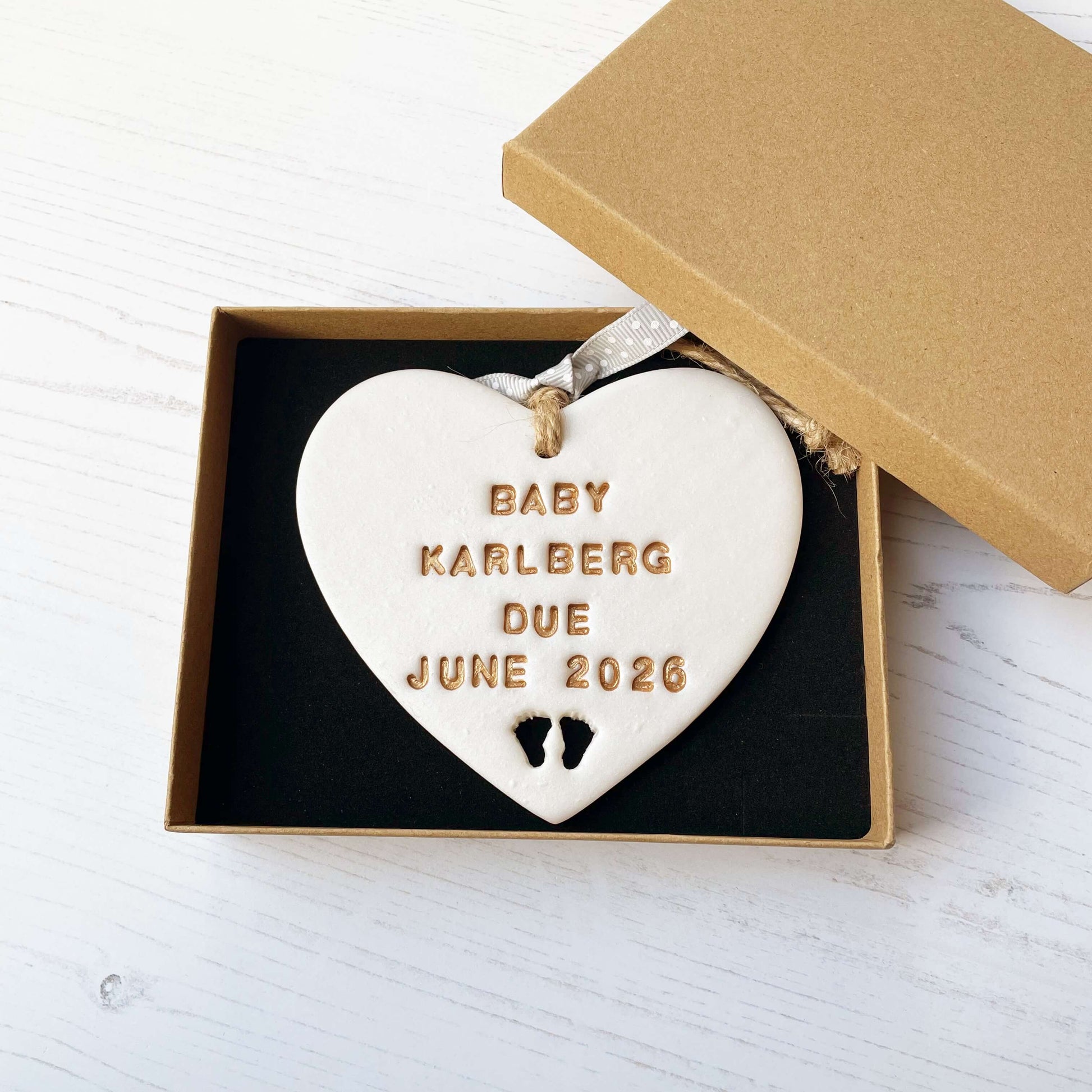 Personalised pregnancy reveal sign keepsake, pearlised white clay hanging heart with baby feet cut out at the bottom, the heart is personalised in gold with BABY KARLBERG DUE JUNE 2026