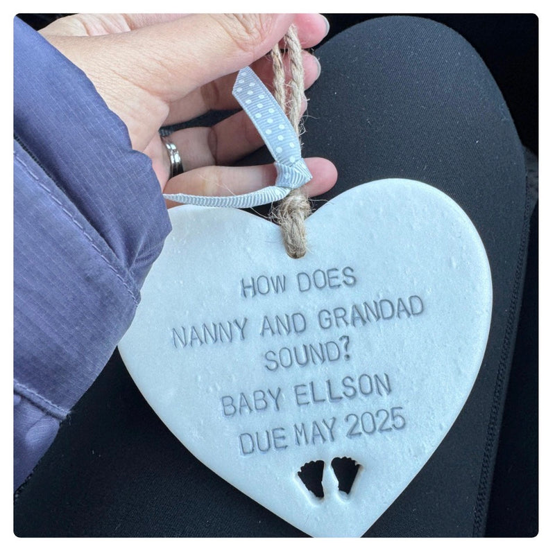 A hand holds a Pearlised white polymer clay heart keepsake hand stamped with the text 'HOW DOES NANNY AND GRANDAD SOUND? BABY ELLSON DUE MAY 2025' and small footprints at the bottom. The plaque hangs from a white ribbon with blue polka dots and a natural twine loop, shown against a dark car seat background.