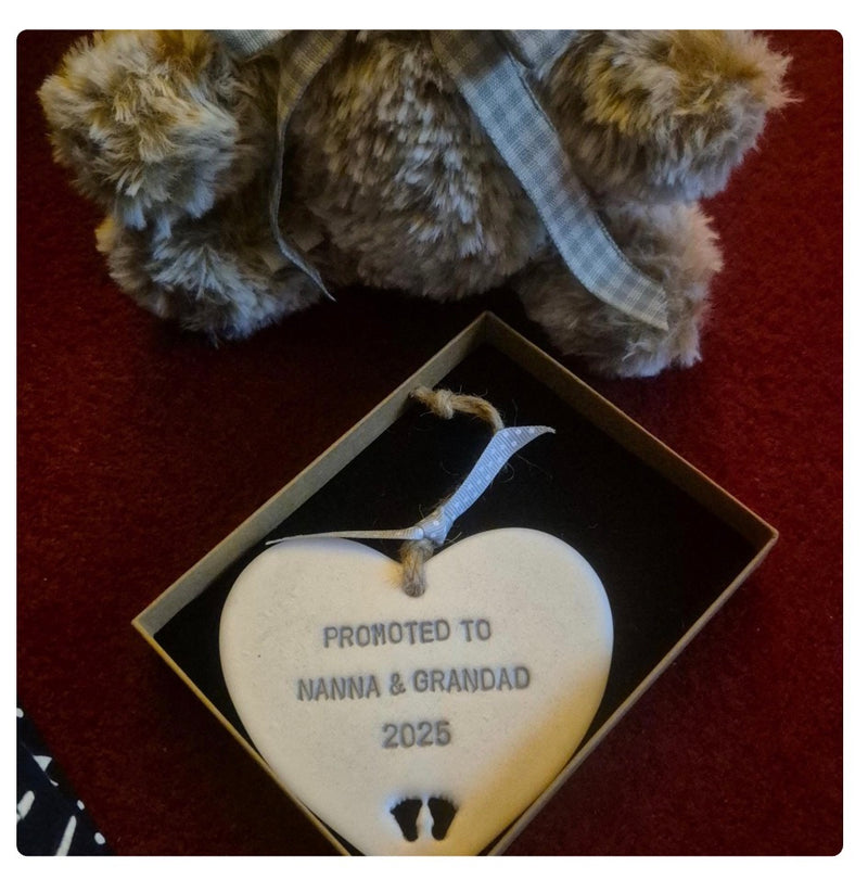 A Pearlised white polymer clay heart inside a gift box, personalised with the text 'PROMOTED TO NANNA & GRANDAD 2025' and small footprints, with a teddy bear partially visible in the background on a red surface.